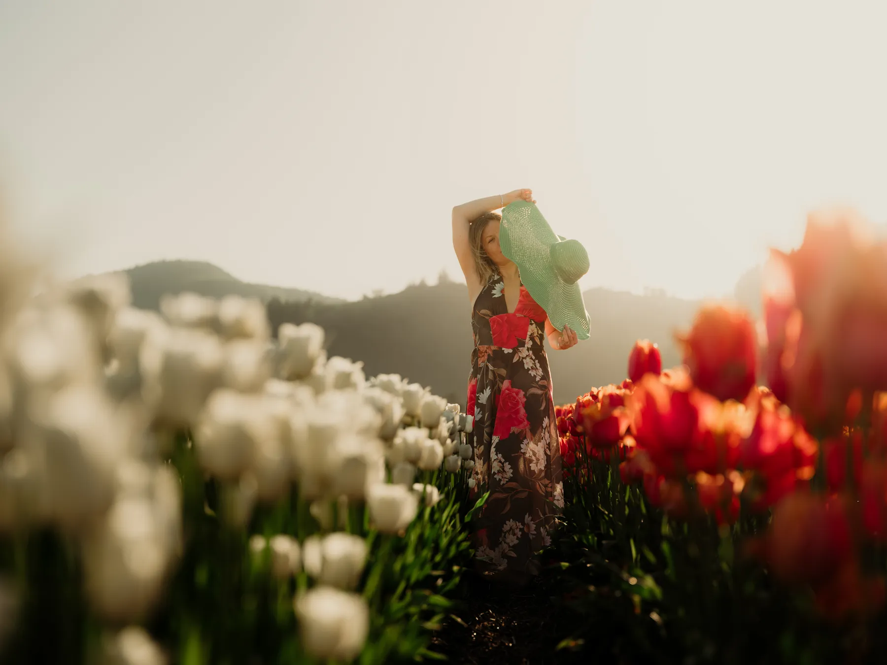 A woman in a floral dress lifts a wide green sun hat between rows of white and red tulips at golden hour, with hills behind her.