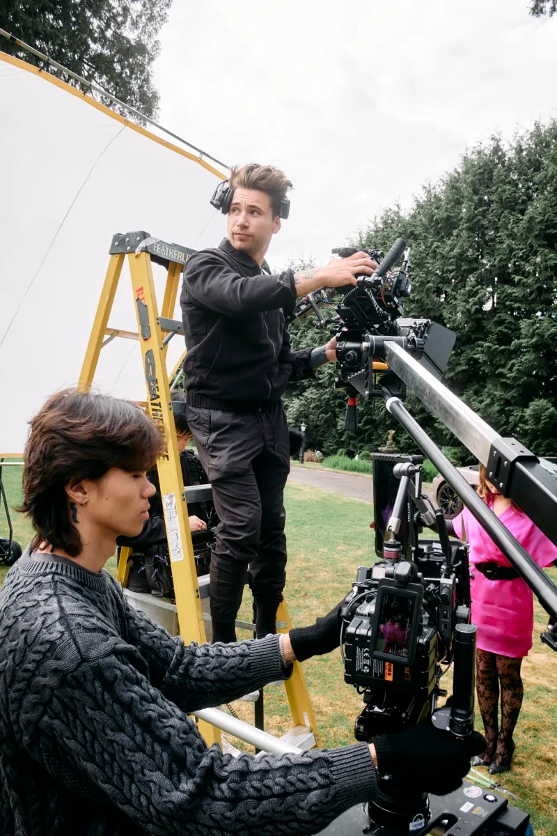 A camera operator on a yellow ladder and a focus puller below work a cinema camera on a jib arm outdoors.
