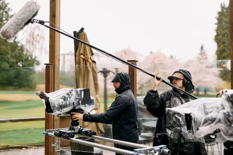 Two crew in rain jackets shoot with a covered cinema camera and a boom microphone on a wet golf-course terrace.