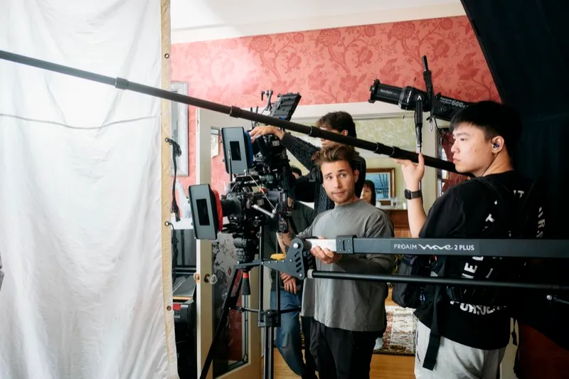 A camera operator and an assistant set a cinema camera beside a white bounce in a room with red patterned wallpaper.