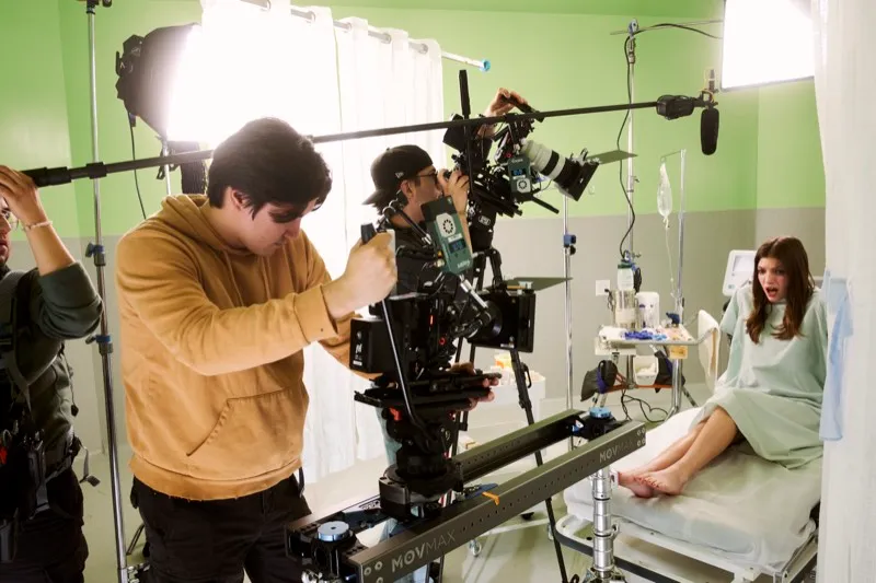 A camera operator frames a shot on a slider in a hospital-room set while an actress in a gown sits on the bed.