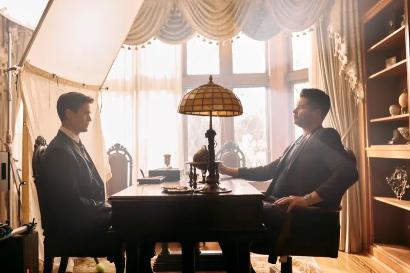 Unit still of two men in suits facing each other across a desk lit by a stained-glass lamp in front of a bright window.