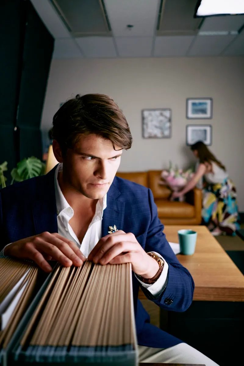 Unit still of a man in a navy blazer leaning over a stack of folders, looking off to the side in an office.