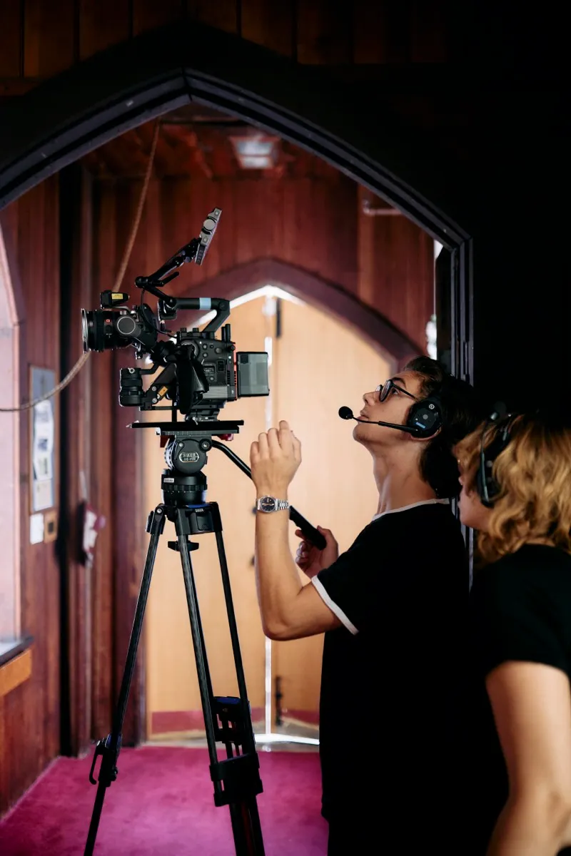 Two crew members set a cinema camera on a tripod in a Gothic wooden doorway with red carpet.