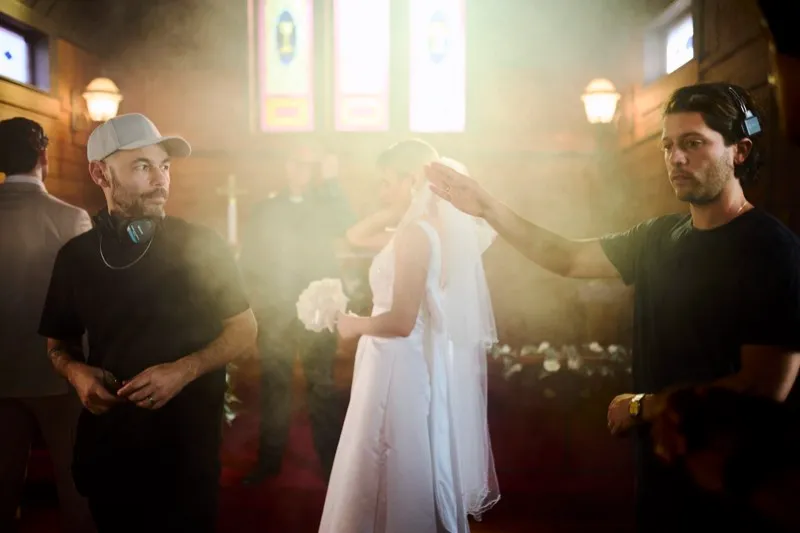 A crew member adjusts a bride's veil on a church set as the director watches from the left, backlit by haze.