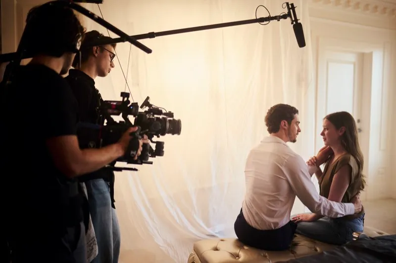 Two crew with a shoulder-mounted camera and boom film a couple sitting together on a bench under a sheer canopy.