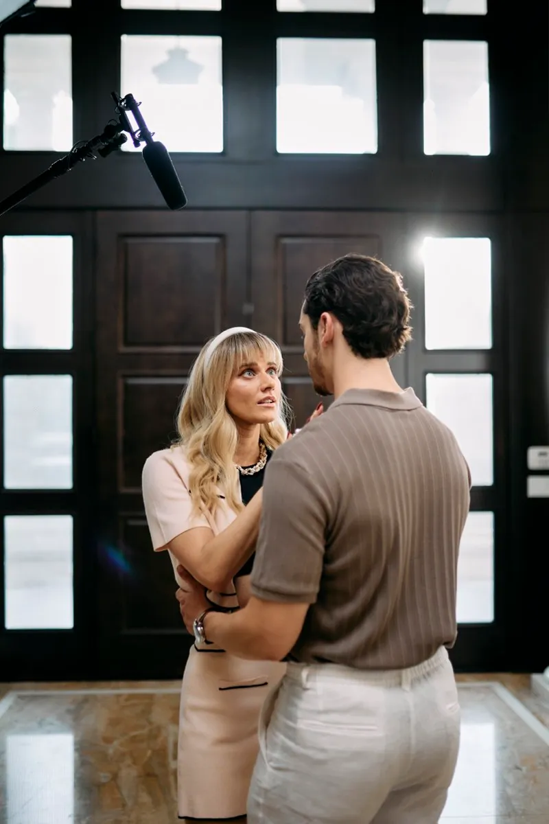 Unit still of a woman in white holding a man's arm in front of tall black doors, a boom microphone above them.
