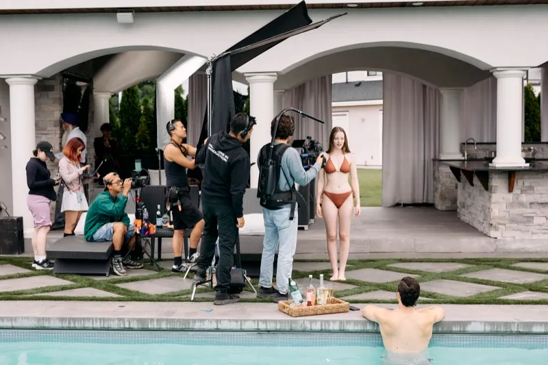 A crew films a woman in a swimsuit beside a pool under a black flag while an actor waits in the water.