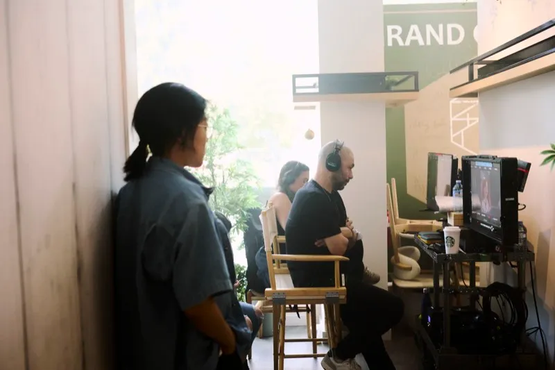 A crew member leans on a doorframe while two people watch playback on a monitor in a sunlit room.