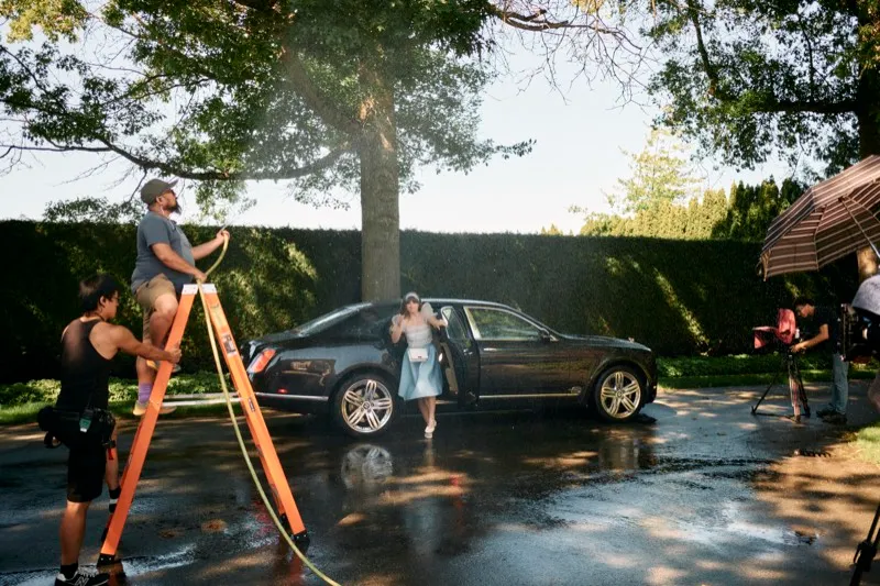 A crew member on an orange ladder holds a light above an actress beside a black car on a wet driveway.