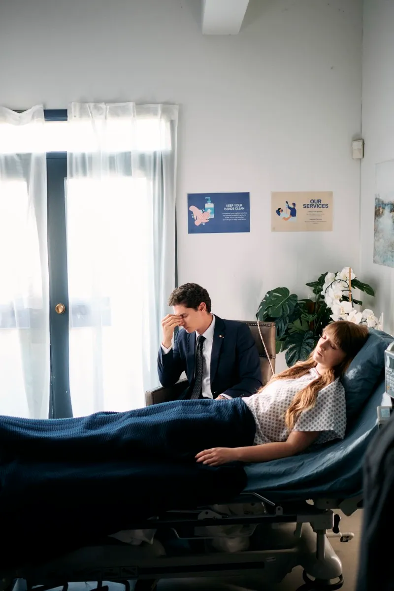 Unit still of a man in a suit holding his head beside a woman lying on a hospital bed by a bright window.
