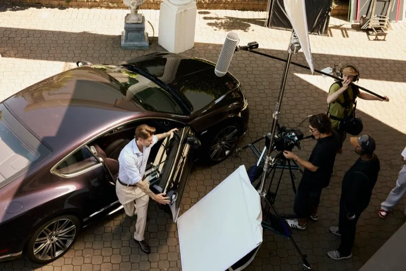 Seen from above, a crew with a boom and a bounce film an actor stepping out of a dark sedan.