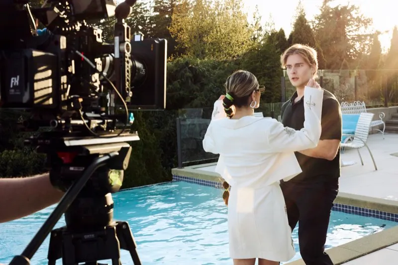 A cinema camera in the foreground films a couple in white beside a swimming pool in low sun.