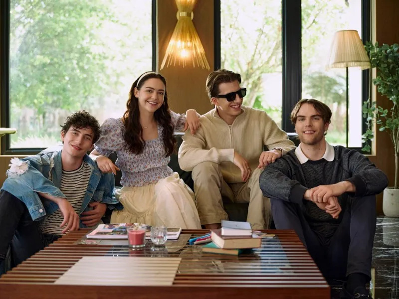 Unit still of four young cast members lounging on a sofa around a coffee table in a warmly lit living room.