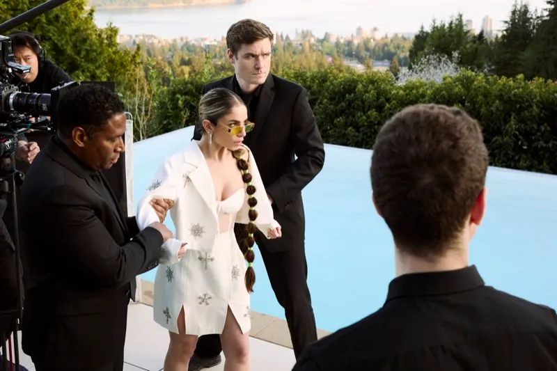 A crew films a woman in a white coat and a man in a black suit beside an infinity pool overlooking the city.