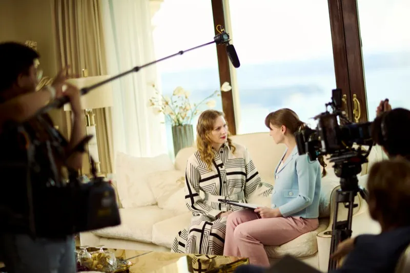 A boom operator and camera film two women talking on a sofa in a bright hotel suite.