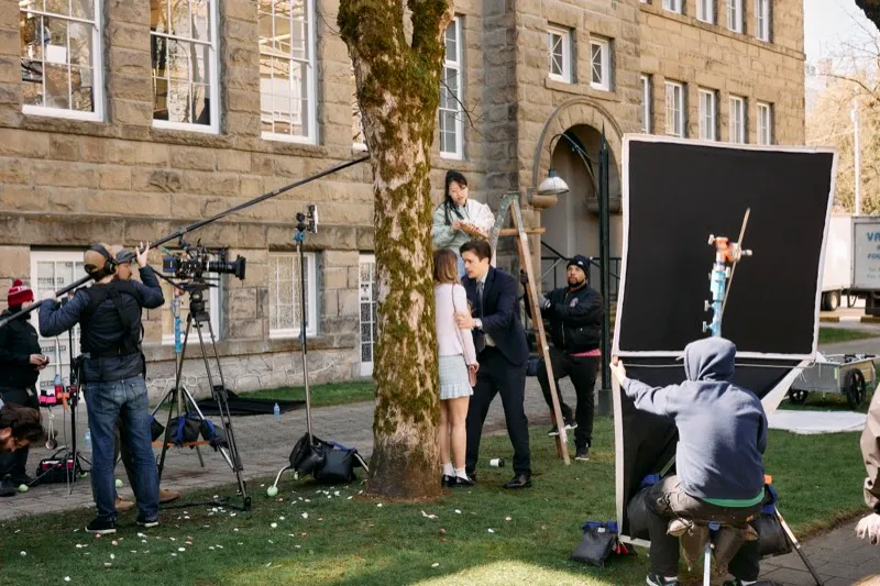 A film crew with a flag and a camera dolly shoot a couple on the lawn in front of a stone school building.