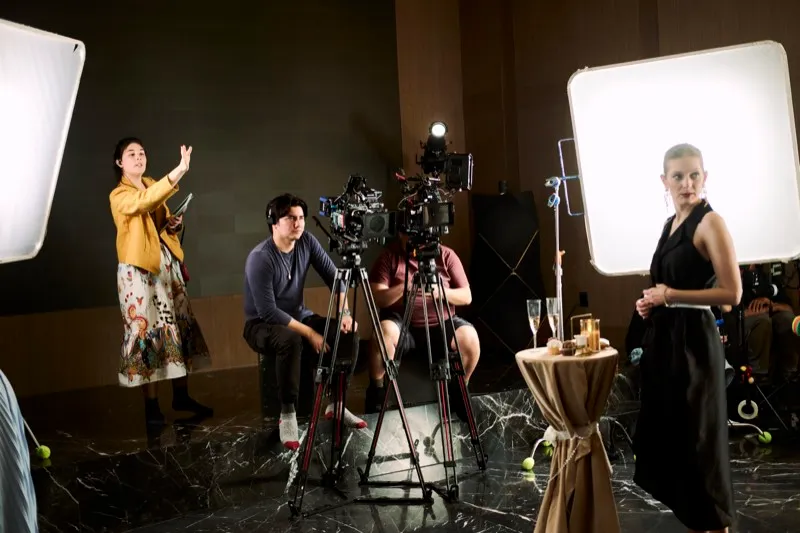 A crew with two soft lights films a woman in a black dress beside a cocktail table in a wood-panelled room.