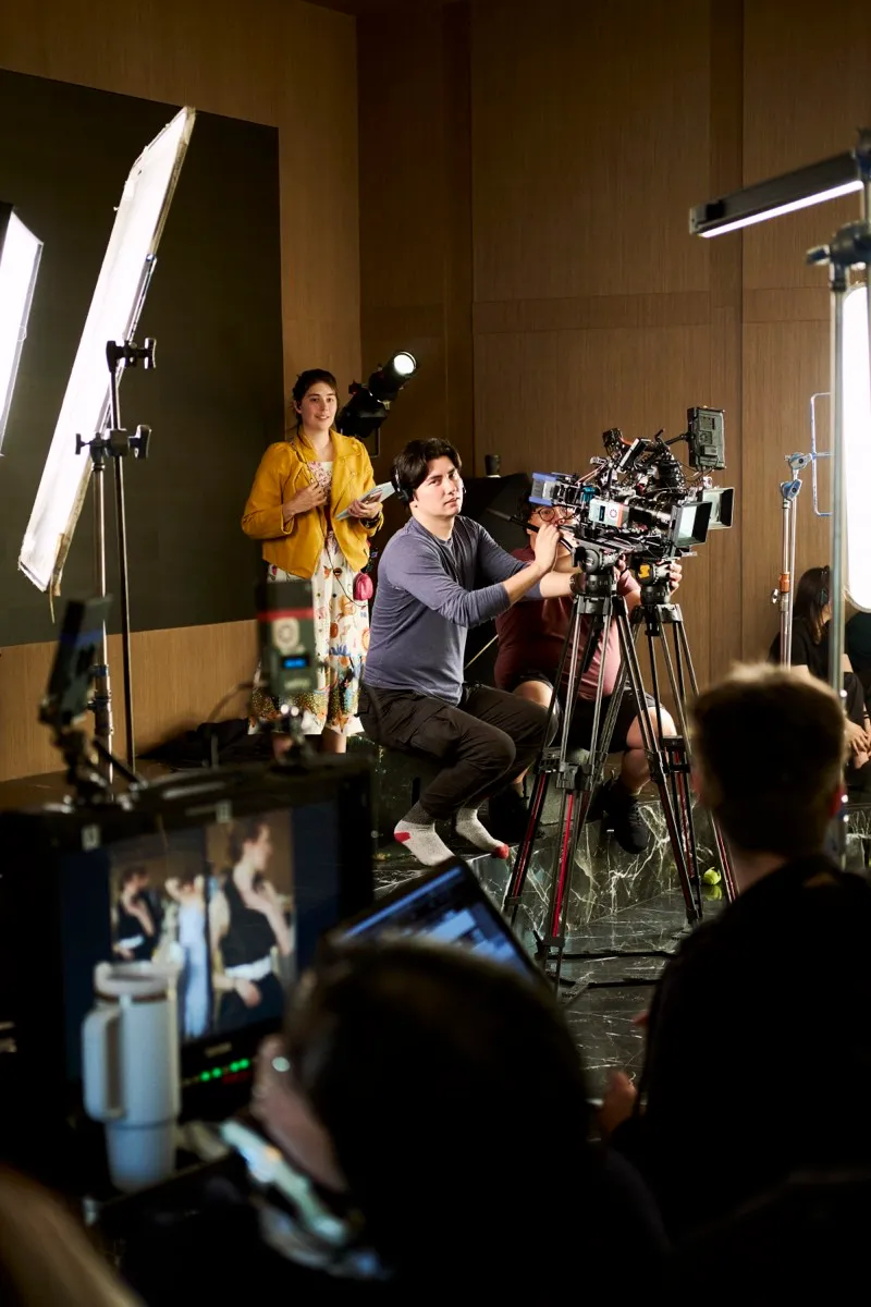 A camera operator seated at a cinema camera films a woman in a yellow cardigan under a large soft light.
