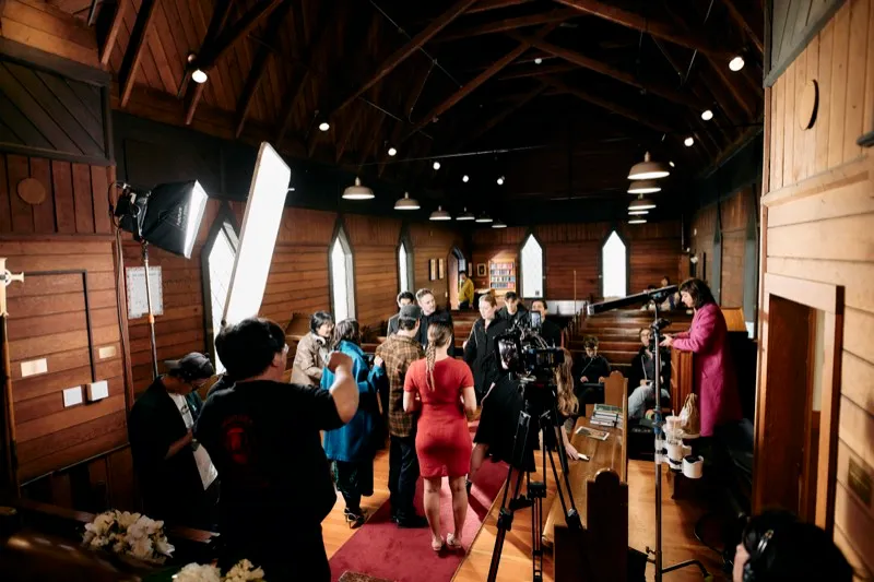 A film crew and cast fill the aisle of a small wooden church between the pews.