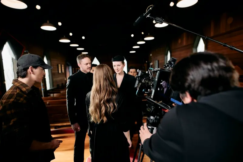 Three actors in dark clothes stand in a church aisle while a camera operator films from the right.