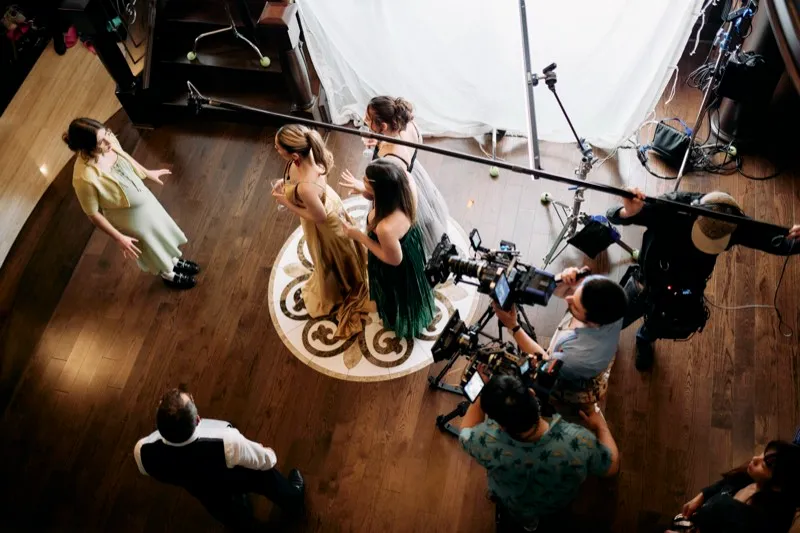 Seen from above, three actresses in gowns are filmed in a circular room by a crew with a camera and boom.