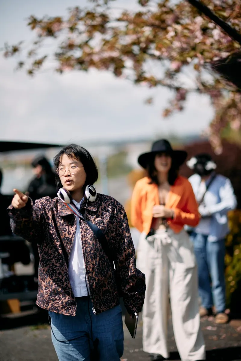 A crew member with headphones round her neck points off frame under cherry blossom, actors waiting behind her.