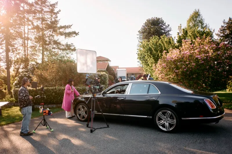 A film crew with a bounce and a camera on a tripod shoot a woman in pink beside a black limousine at sunset.