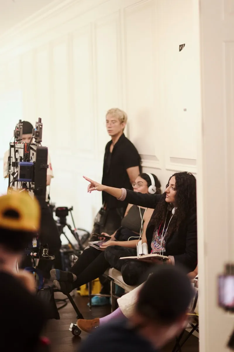 A director with dark curly hair points across the set from her chair while crew work the camera beside her.