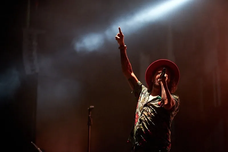 A singer in a wide-brimmed hat points to the sky as a spotlight beam cuts through the haze above him.