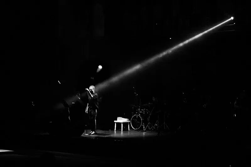 Black and white photograph of a lone singer in a single spotlight on an otherwise dark stage.