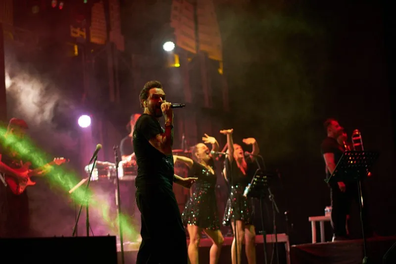 A singer with two backing vocalists in sequinned dresses under green and pink haze.