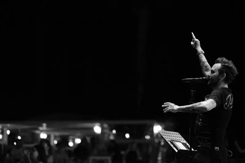 A tattooed singer points to the sky at the front of a stage, the crowd small in the dark behind him.
