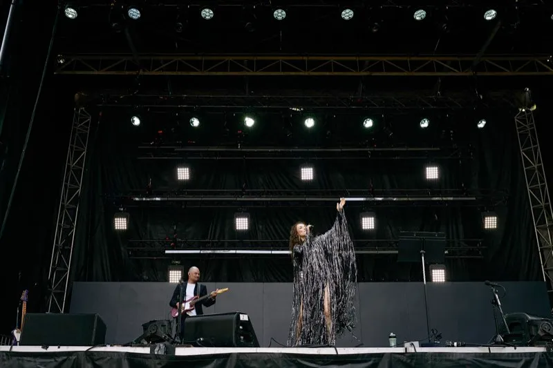 A singer in a silver fringed dress raises her arm on an outdoor festival stage beside a guitarist.
