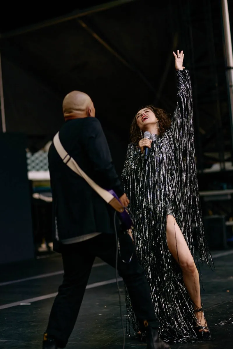 A singer in a silver fringed dress throws her head back beside a guitarist on a festival stage.