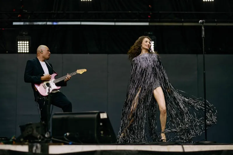 A singer in a silver fringed dress and a guitarist with a pink guitar perform on an outdoor stage.