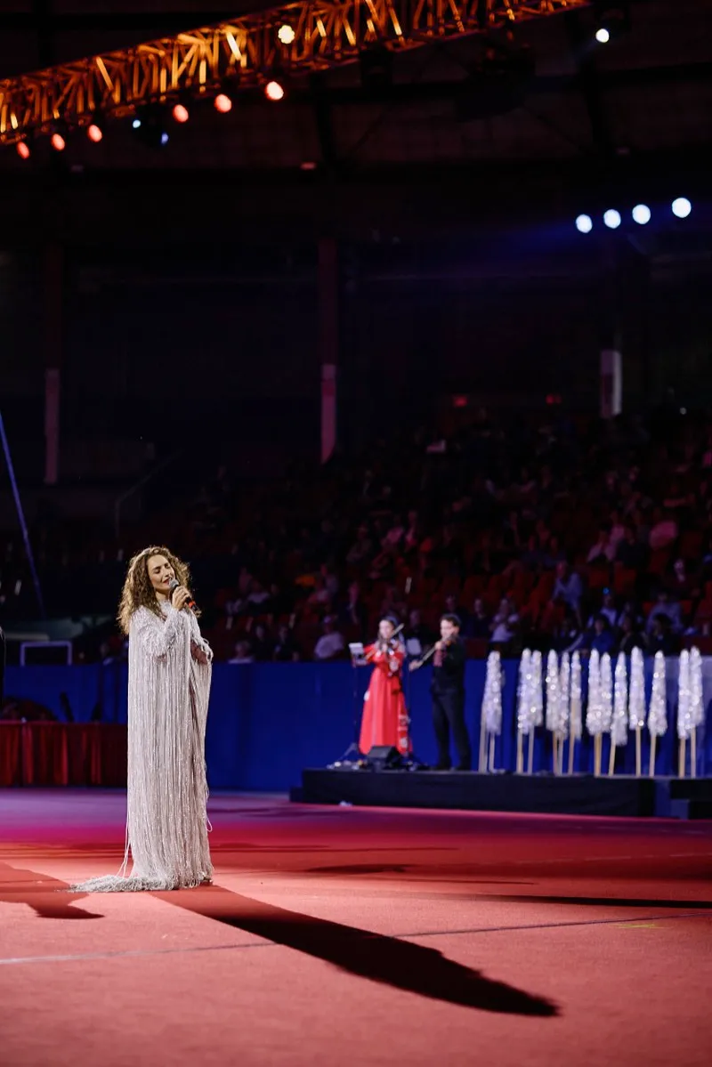 A singer in a long white beaded gown performs on a red arena floor with a military band and flags behind her.