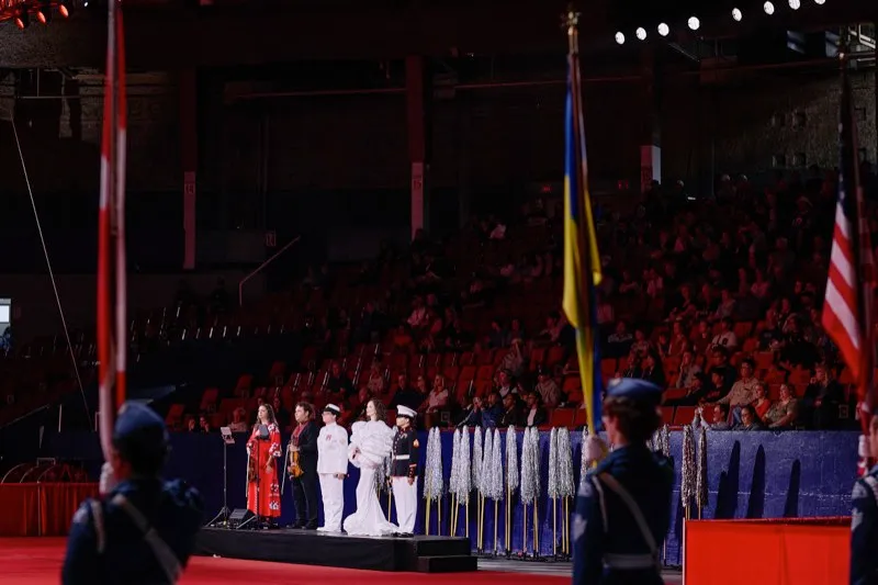 Performers in red and white stand on a platform in an arena, flag bearers in the foreground.