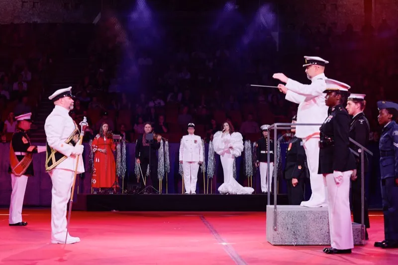 Naval officers in white uniforms on an arena floor, a conductor raising his baton on a platform behind them.