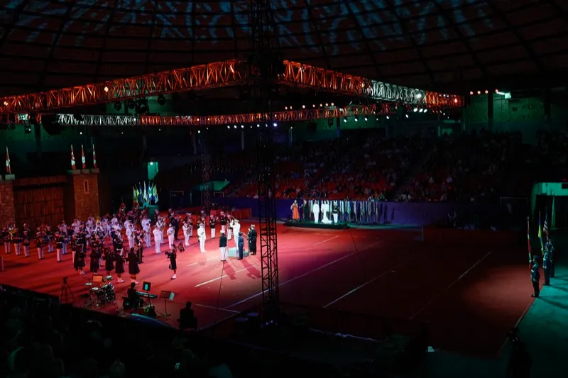 A wide view of a military tattoo in an arena, pipers and drummers on the red floor and the crowd in the stands.