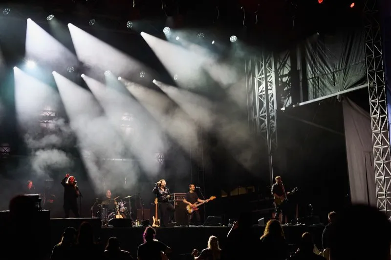 A band on an outdoor stage under white beams cutting through haze, the crowd in front.