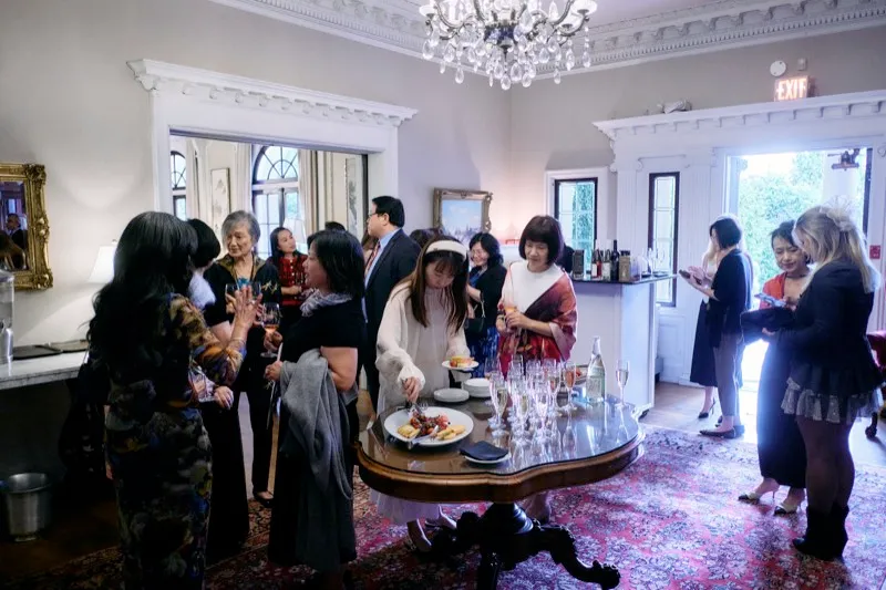 Guests mingle around a table of glasses and hors d'oeuvres in a panelled reception room under a chandelier.