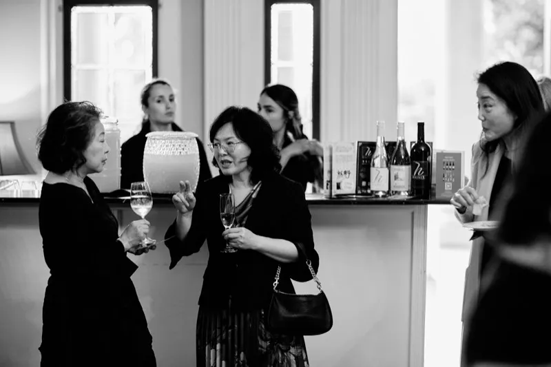 Black and white photograph of guests talking over wine glasses beside a bar in a bright reception room.