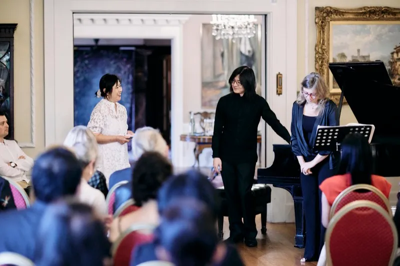 A host addresses seated guests beside a grand piano at a private recital.