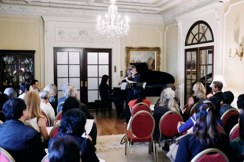 Guests seated on red chairs listen to a flautist and pianist beneath a chandelier in a period drawing room.