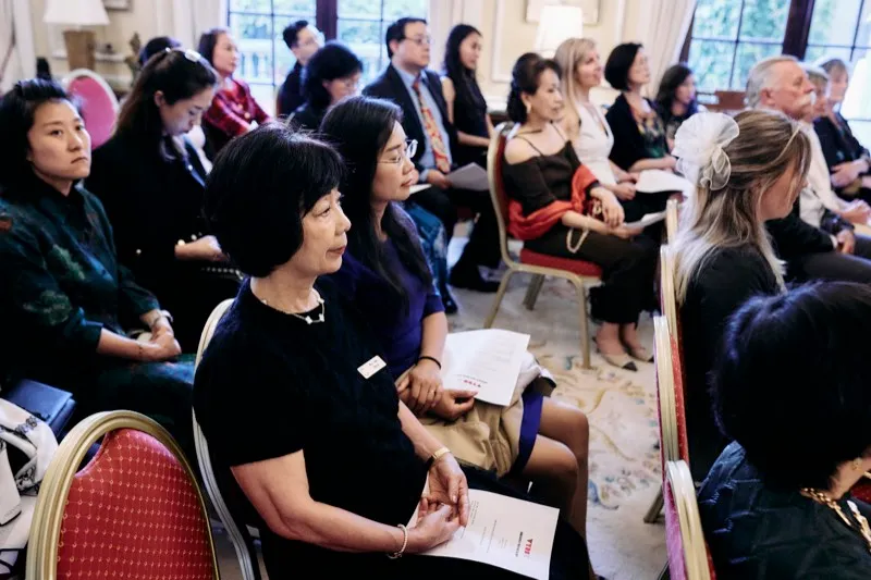 Guests in evening dress listen from their seats, one holding a printed programme.