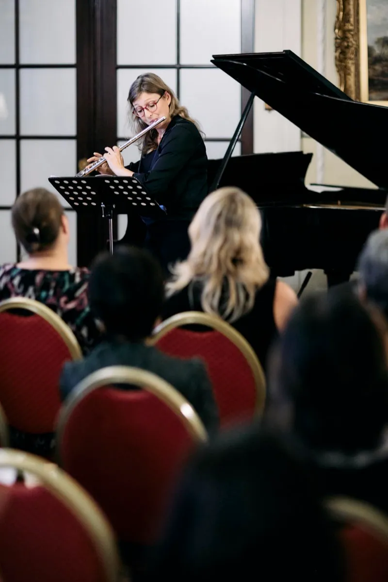 A flautist plays beside a grand piano, seen over the heads of the seated audience.