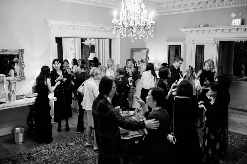 Black and white photograph of a reception crowd under a chandelier, one guest greeting another with a hand on her arm.