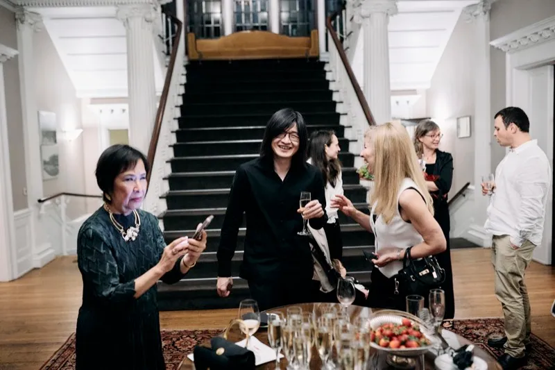 Guests talk on the landing of a grand staircase, a table of champagne and strawberries in the foreground.