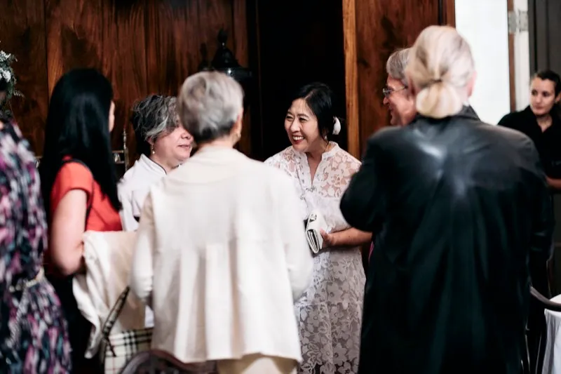 Guests in conversation in a wood-panelled room, one in a white lace dress.
